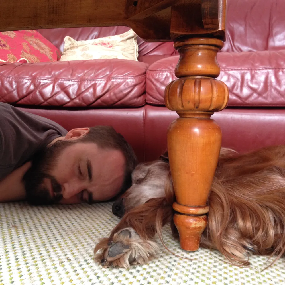 Image contains a photo of a man laying on the floor with their head against the head of a dog who is also laying on the floor.
